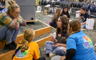 A group of people sit around on the floor and talk to each other