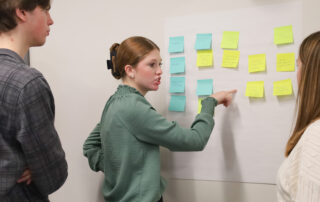 Picture of a young woman pointing to sticky notes hanging on a wall while talking to another student.