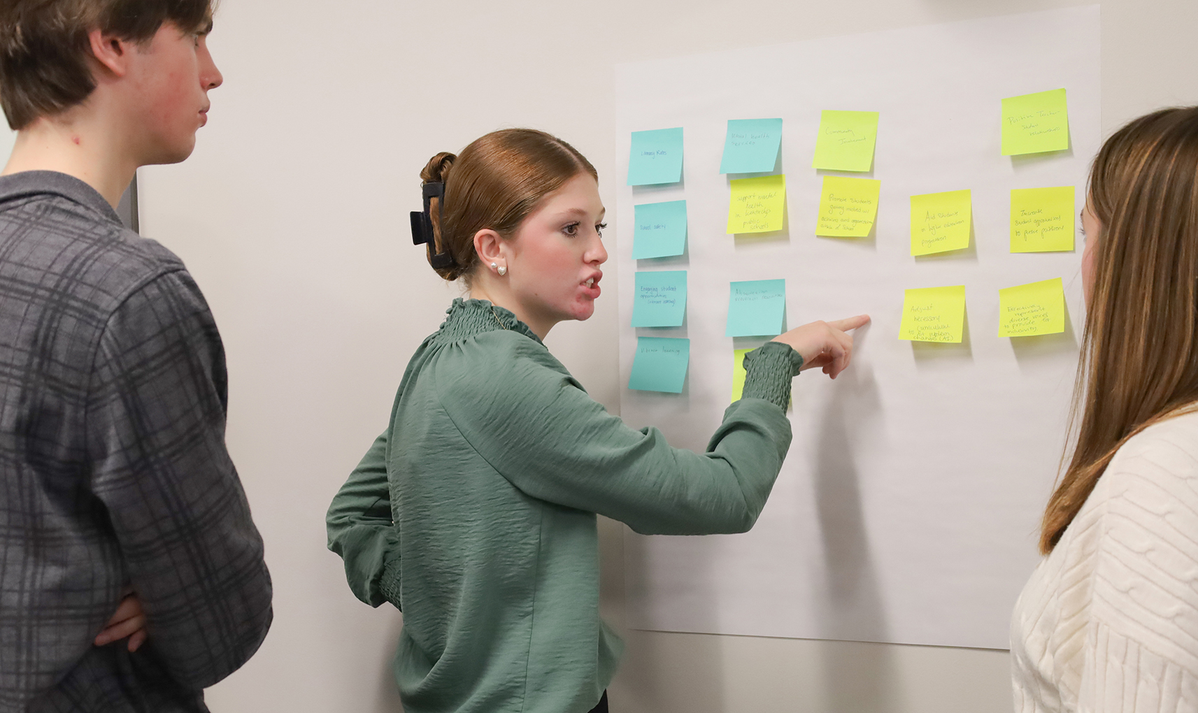 Picture of a young woman pointing to sticky notes hanging on a wall while talking to another student.