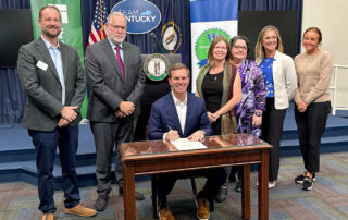 A group of people stand around Gov. Andy Beshear, who is seated at a table, holding a pen to paper while smiling for a posed photo