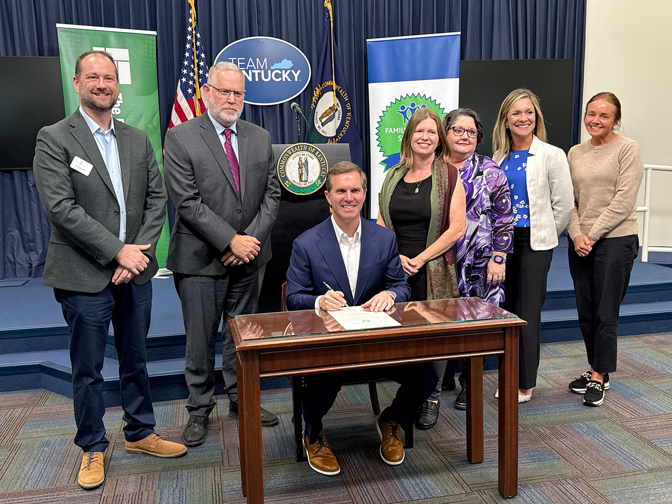 A group of people stand around Gov. Andy Beshear, who is seated at a table, holding a pen to paper while smiling for a posed photo