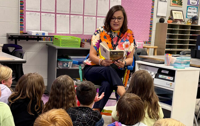 Michele Bradley sits in a chair and reads a book to a classroom full of kids sitting on the floor in front of her
