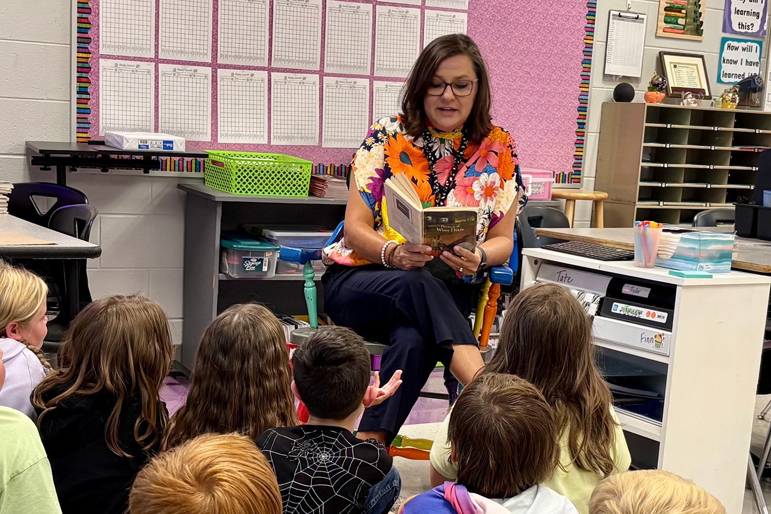 Michele Bradley sits in a chair and reads a book to a classroom full of kids sitting on the floor in front of her