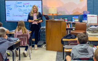 A woman holds a book while standing in front of a classroom of kids who are seated at their desks