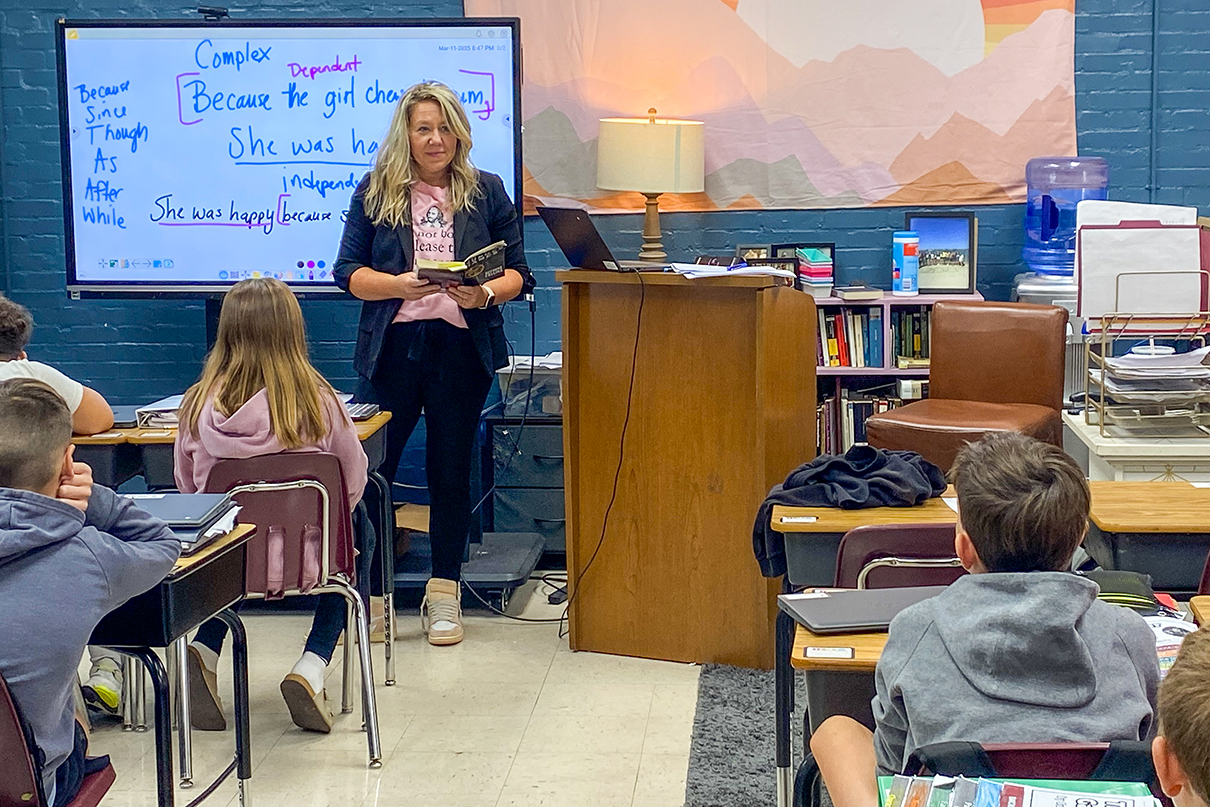 A woman holds a book while standing in front of a classroom of kids who are seated at their desks