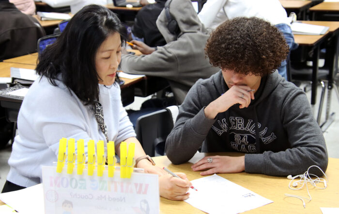 a woman sits down with a student, putting a red pen to a piece of paper