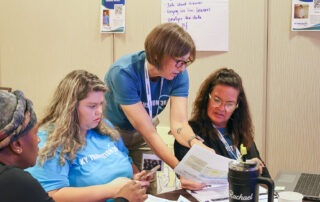 A woman points to a piece of paper as two women who are sitting look at the paper