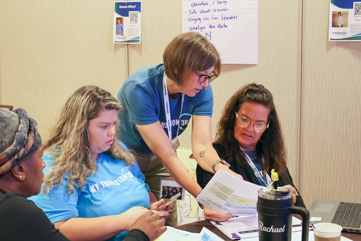 A woman points to a piece of paper as two women who are sitting look at the paper