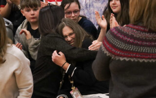 Two people hug amid a crowd of others on the bleachers of a gymnasium
