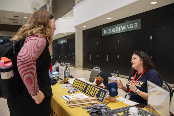 A woman wearing a backpack stands next to a table, talking to a woman who is sitting. A nameplate saying "Murray State" is on the table.