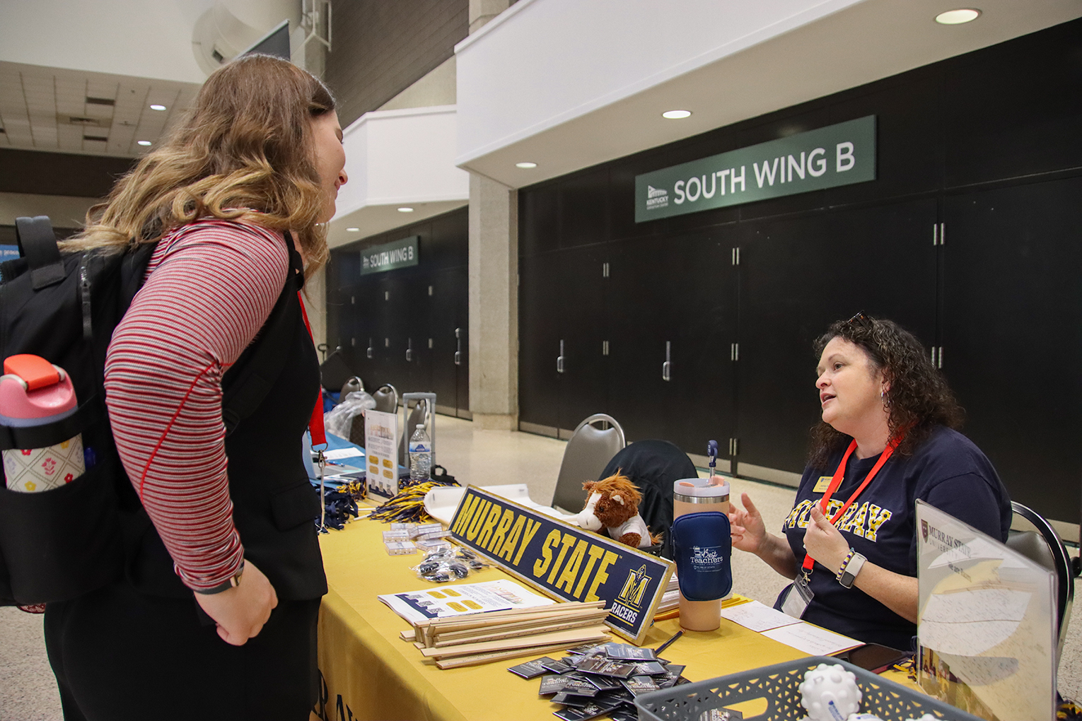 A woman wearing a backpack stands next to a table, talking to a woman who is sitting. A nameplate saying "Murray State" is on the table.