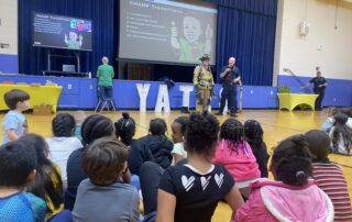 Two firefighters, including one wearing full gear, stand in a gymnasium in front of several kids who are seated on the ground