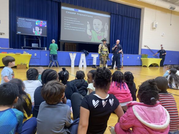 Two firefighters, including one wearing full gear, stand in a gymnasium in front of several kids who are seated on the ground