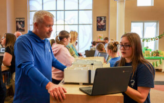A girl makes a presentation to a man in a library setting, both of them looking at a laptop screen as the girl is talking