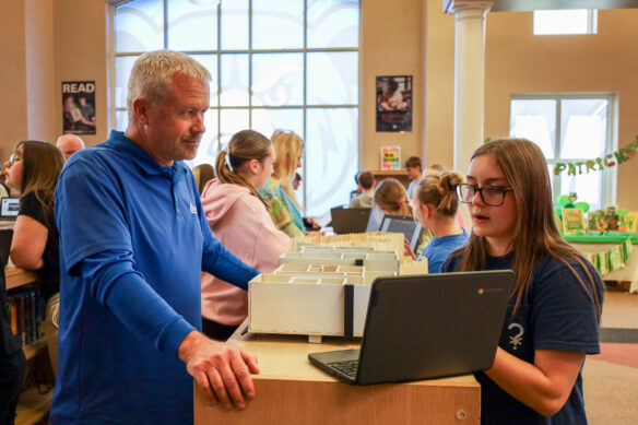 A girl makes a presentation to a man in a library setting, both of them looking at a laptop screen as the girl is talking