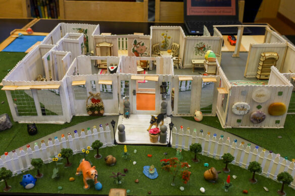 A model home is show, made up of popsicle sticks and small decorative items, with the top off so you can see the floorplan of the home