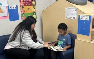 A woman sits with a child in a classroom and helps him read a book