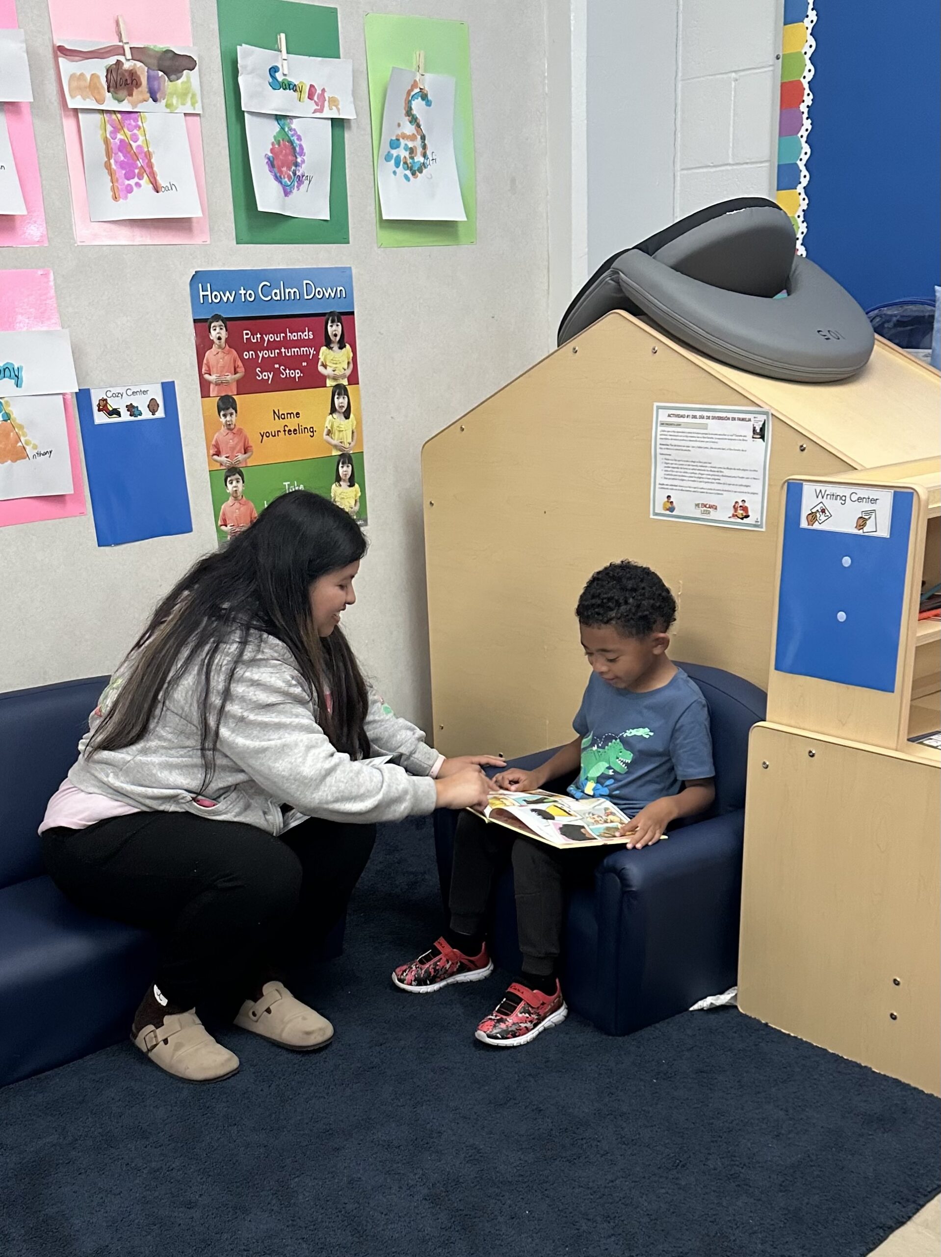 A woman sits with a child in a classroom and helps him read a book