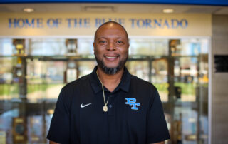 A man stands in front of a school with the words "Home of the blue tornado" behind him