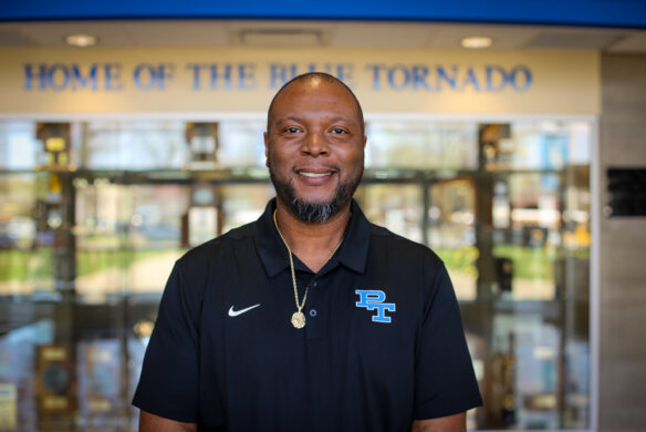 A man stands in front of a school with the words "Home of the blue tornado" behind him