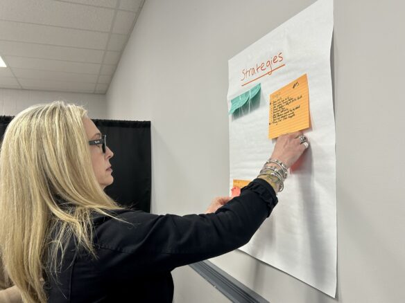 A woman puts a Post It note onto a piece of paper place on a wall that says "Strategies"