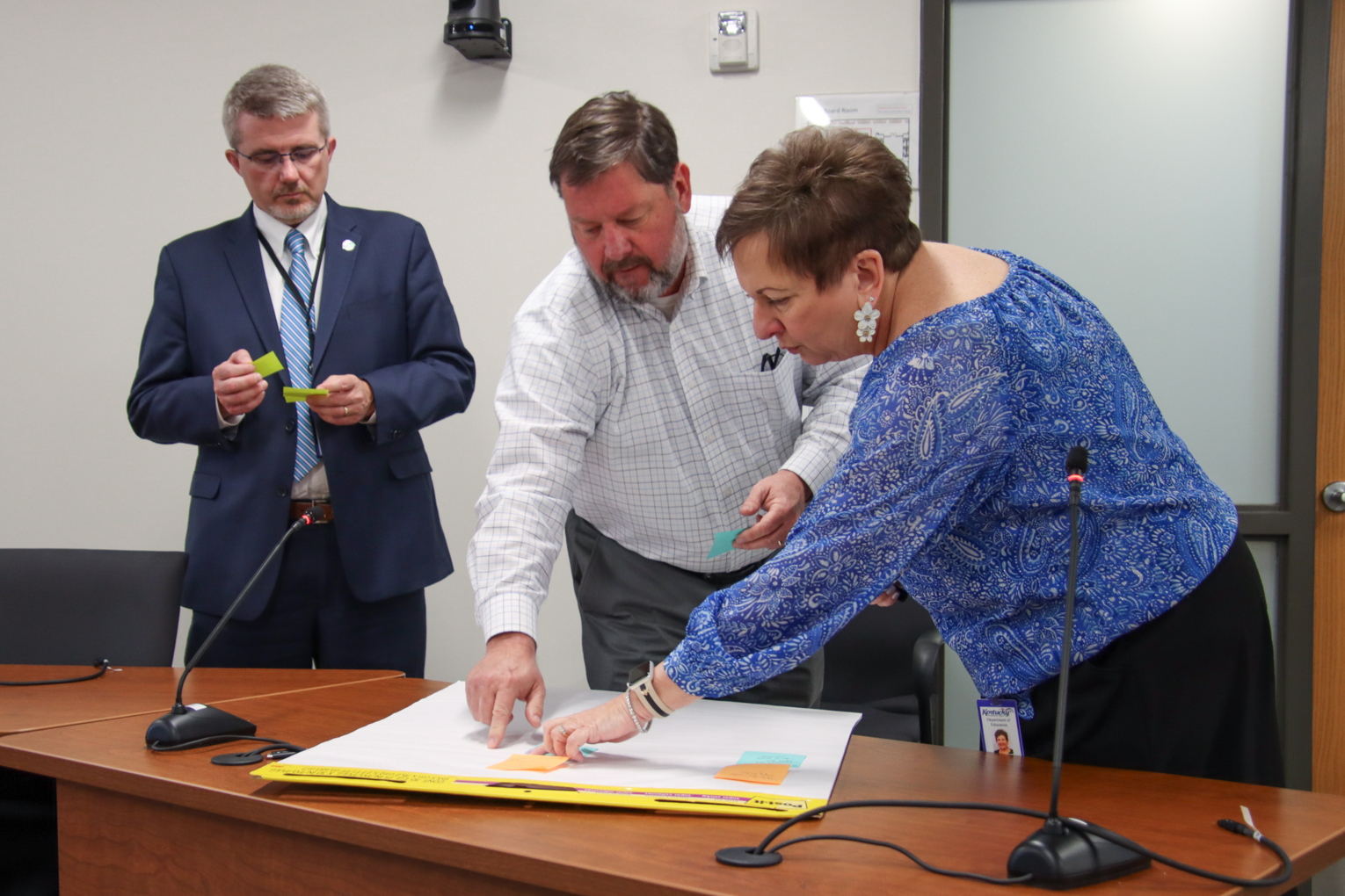 Two people put post it notes on a white piece of construction paper as a third person looks on