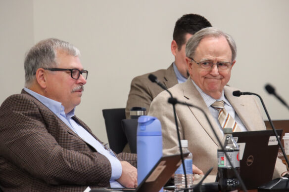 Two men sit next to each other as the man on the right smiles moderately