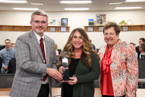 Three people pose for a photo as the woman in the middle and the man on the left hold an award