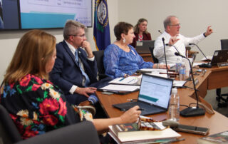 A man holds his hands out demonstratively as three others sitting next to him and another woman sitting behind him look at him