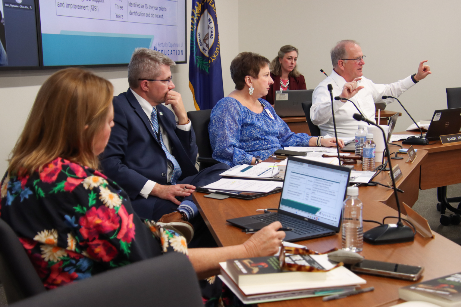 A man holds his hands out demonstratively as three others sitting next to him and another woman sitting behind him look at him
