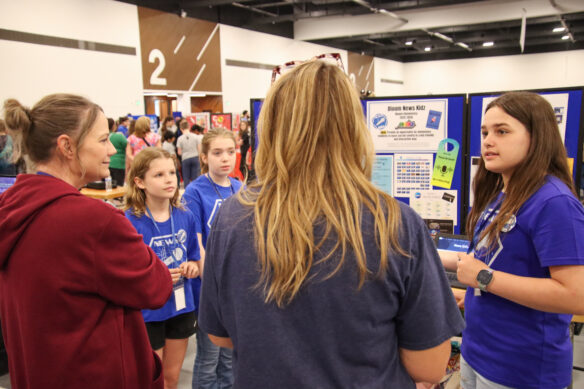 A group of kids present a project to two judges with a poster board behind them, including information about their project