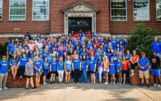 Photo shows a group of dozens of people posing for a large group shot in front of a school building, most of whom are wearing blue shirts