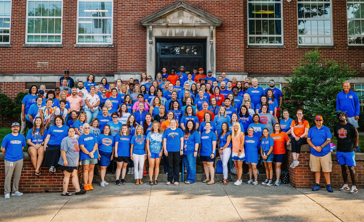 Photo shows a group of dozens of people posing for a large group shot in front of a school building, most of whom are wearing blue shirts