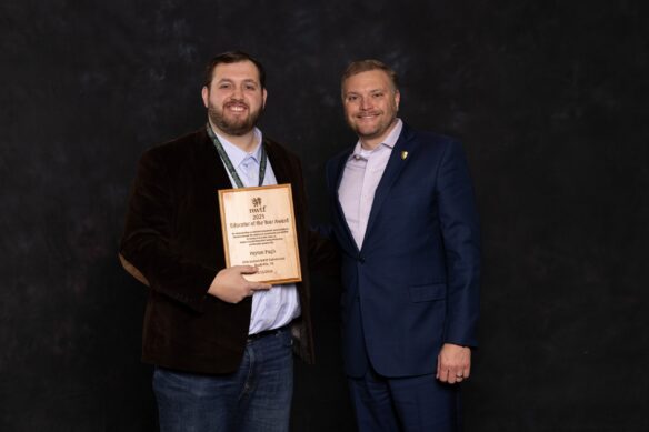 Two men pose for a photo in front of a black backdrop. One man holds an award