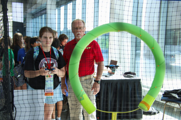A girl holds a remote control and flies a drone through a green hoop as a man in a red polo supervises