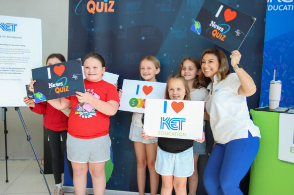 A group of kids pose for a photo with a woman, holding up signs saying they love KET, News Quiz and STLP