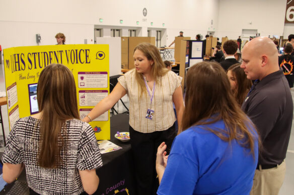 A student points to a poster board and talks to a couple of judges