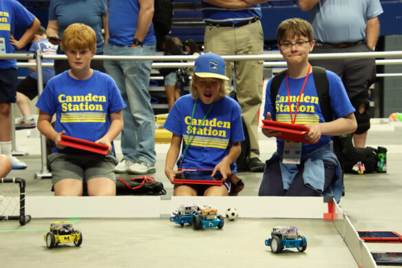 Three students holds tablets and watch robotic cars play with a soccer ball
