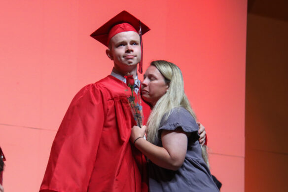 A man in a red graduation cap and gown hugs a woman