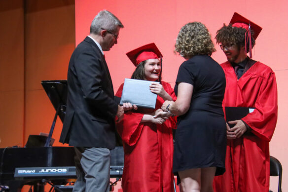 A woman in a red graduation outfit shakes hands with two people as one hands them a diploma