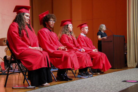 Four people in red graduation caps and gowns sit on a stage as a woman talks into a microphone on a podium