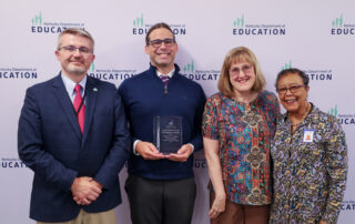 Four individuals posing for a picture in front of a background. Person in middle is holding an award.