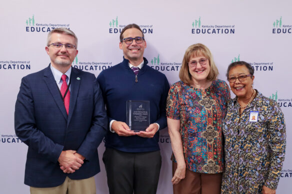 Four individuals posing for a picture in front of a background. Person in middle is holding an award.