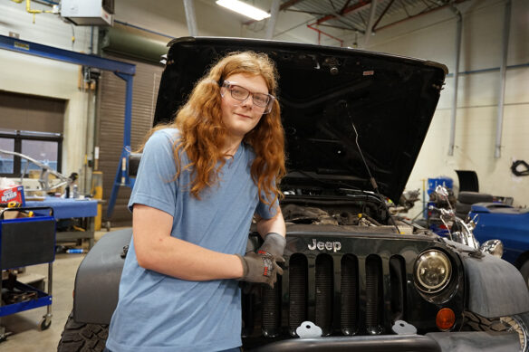 A man with long red hair stands in front of a Jeep Wrangler with the hood up and his arm casually resting on the front grill of the vehicle.
