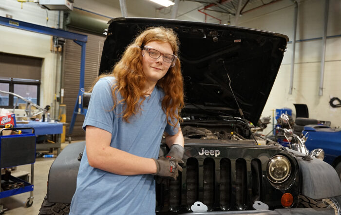 A man with long red hair stands in front of a Jeep Wrangler with the hood up and his arm casually resting on the front grill of the vehicle.