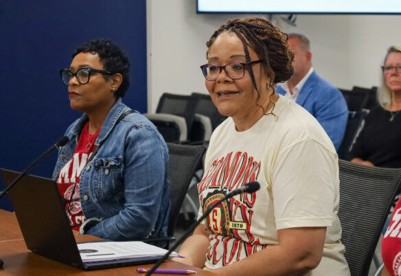 Two women sit at a table as one speaks into a microphone