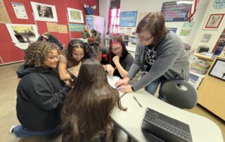 A woman stands over a table, pointing to a piece of paper as students sit around the table