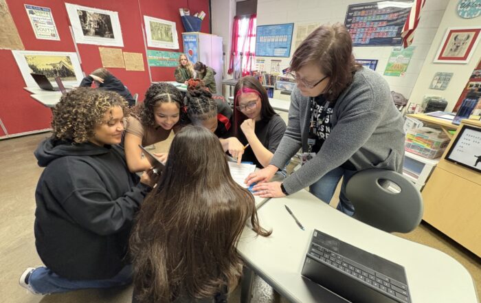 A woman stands over a table, pointing to a piece of paper as students sit around the table