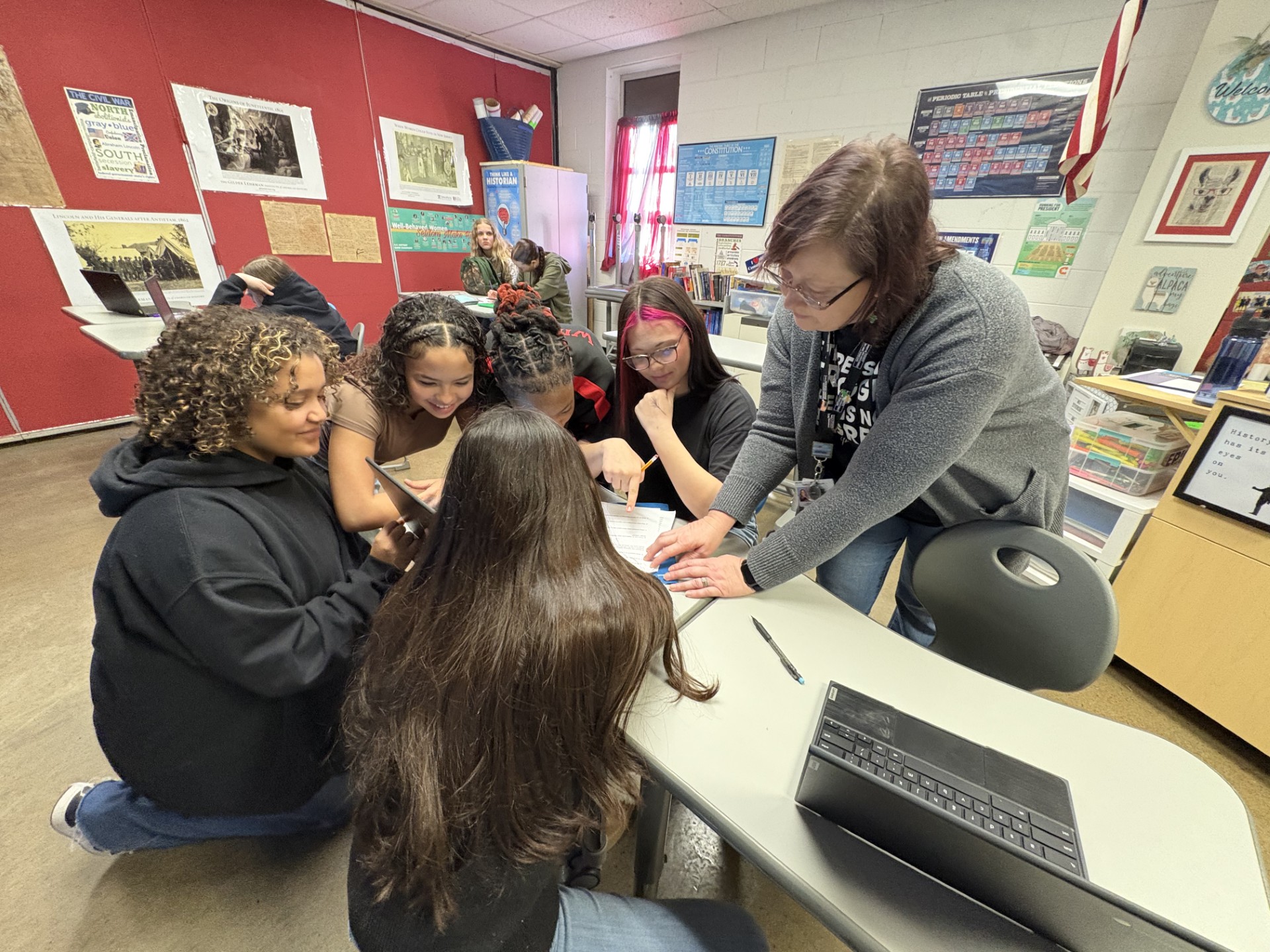 A woman stands over a table, pointing to a piece of paper as students sit around the table