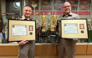 Tom Krupp and Larry Lee pose in front of an awards case holding large framed certificates and statues.